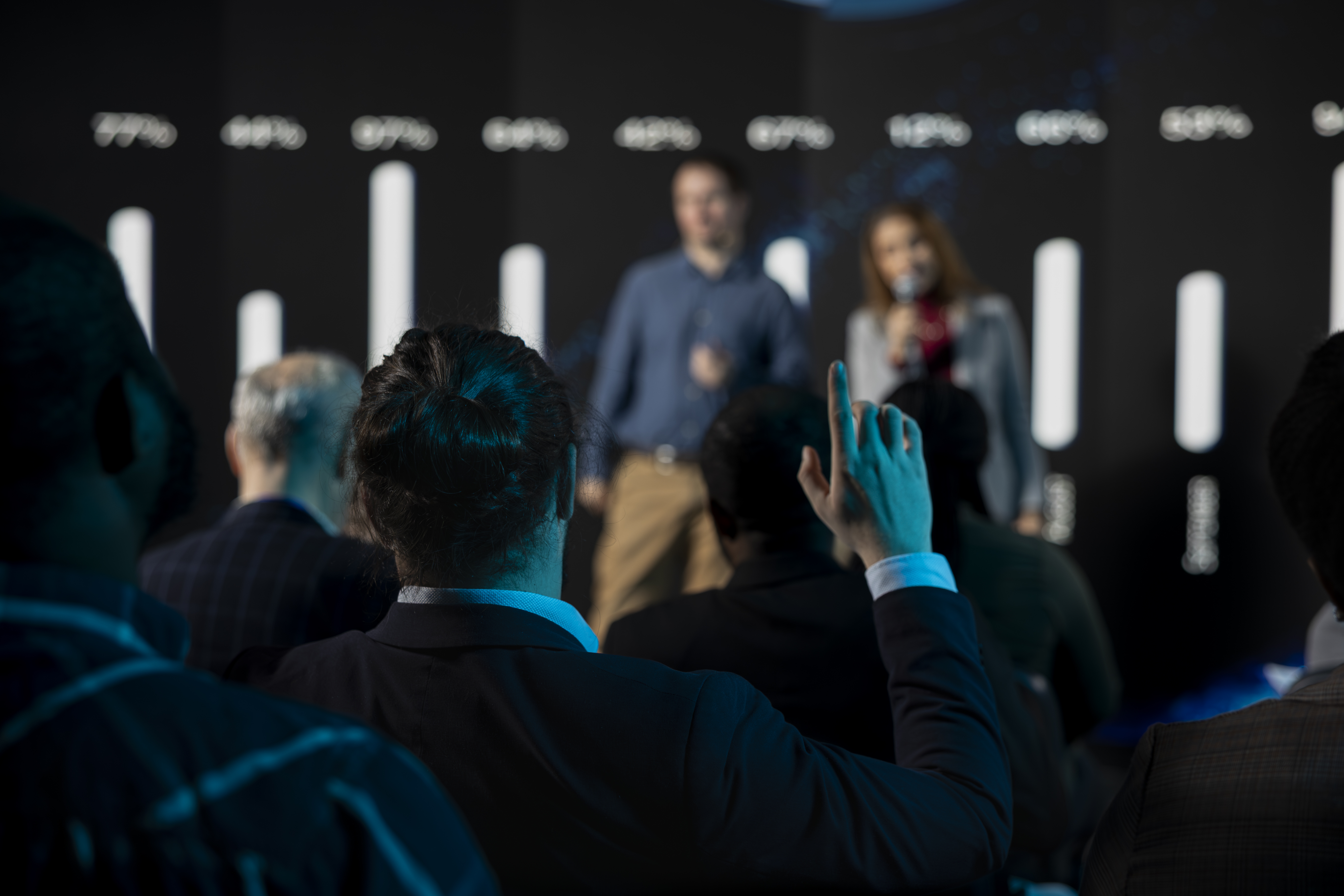 Male listener in audience asking questions during presentation and panel discussion, public speakers team engaging in a debate with attendees. Raising hand and being active in conference hall.