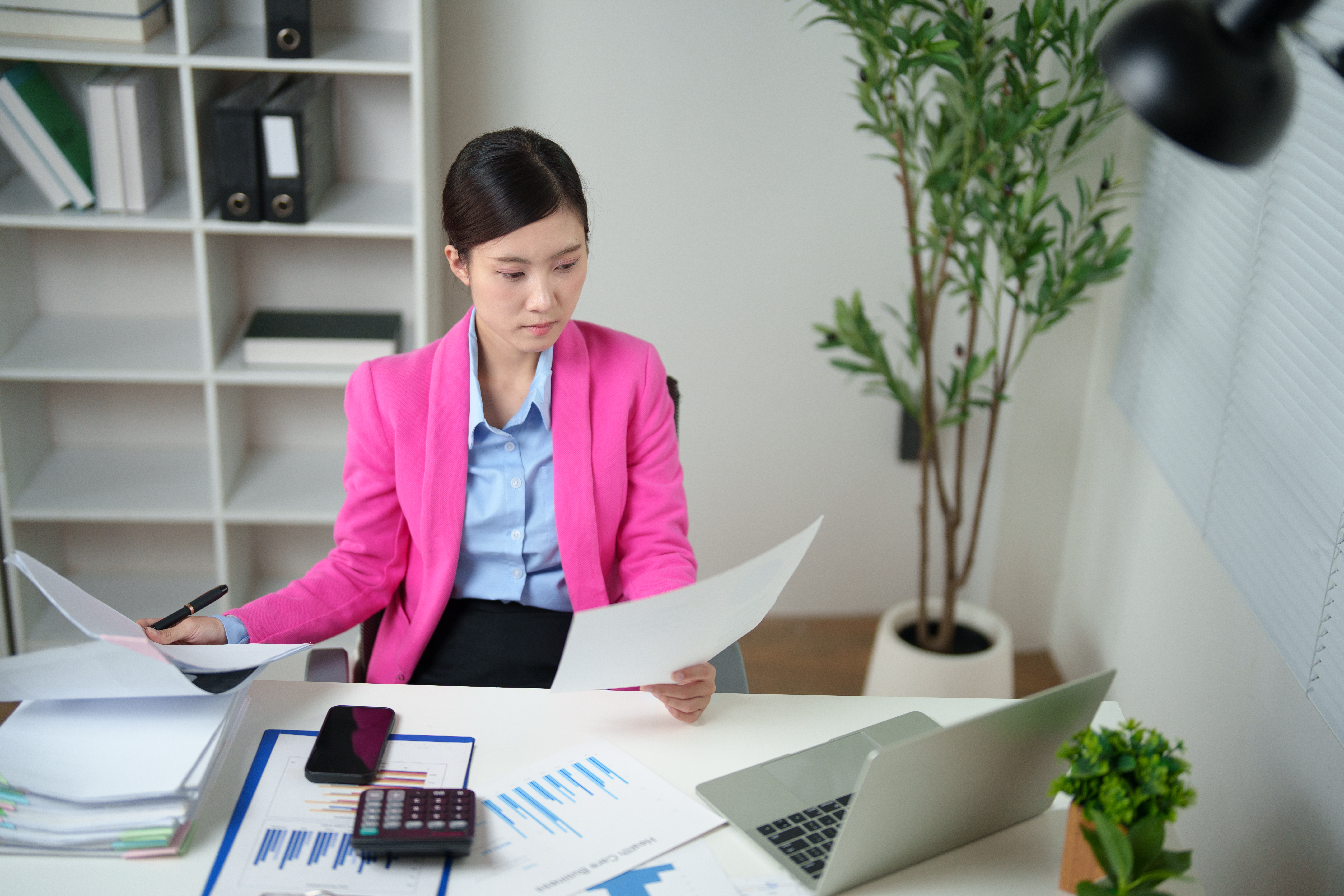 Young adult Asian businesswoman wearing a pink blazer, concentrating on reviewing financial documents and paperwork at her modern office desk during a workday, analyzing data and managing reports