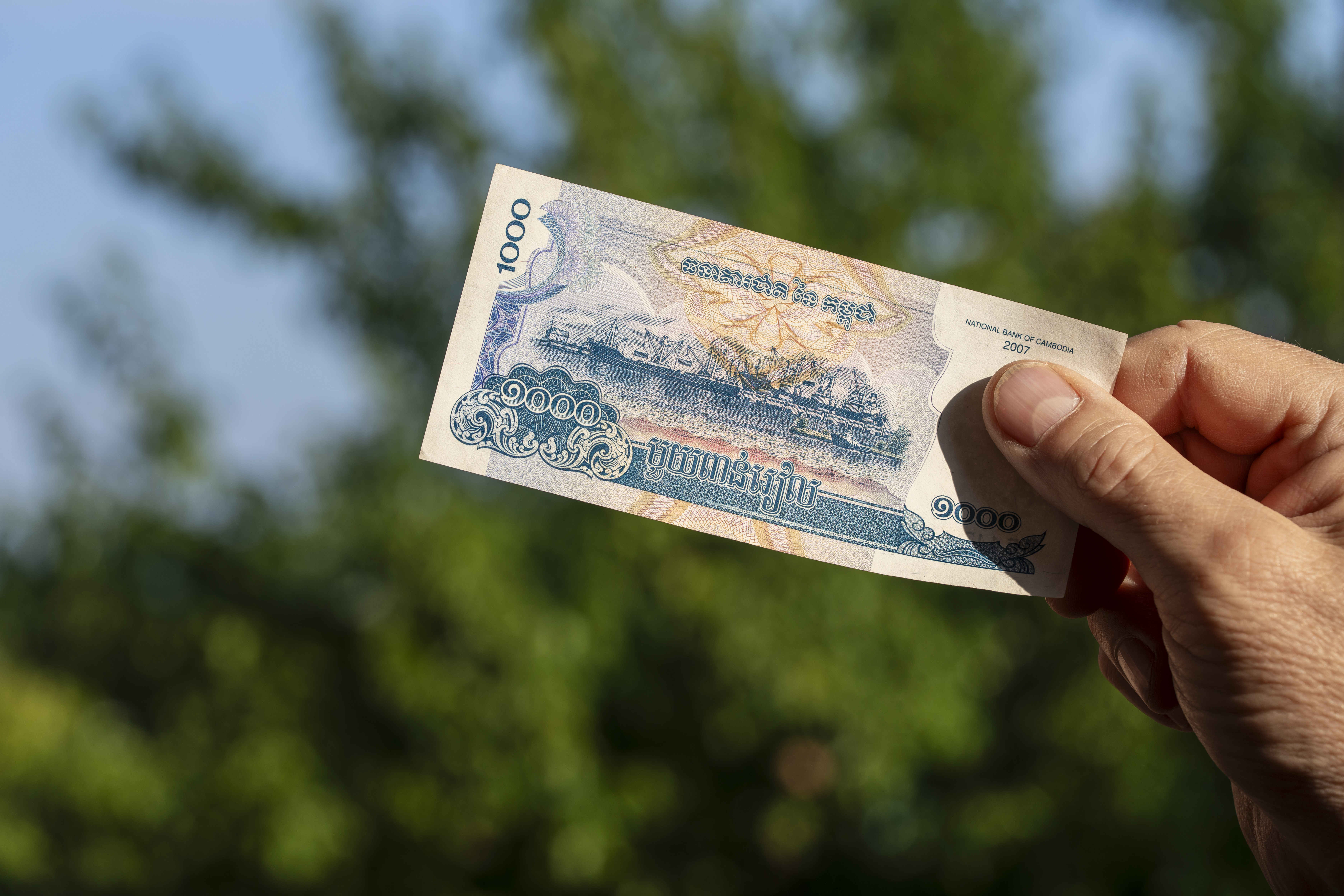 Male hand holding Cambodian riel banknote outdoors. Close up of person holding 1000 Cambodian riel cash bill issued by National Bank of Cambodia against blurred green trees backdrop